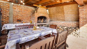 Un espacio comedor al aire libre con horno de ladrillo, mobiliario de madera y una mesa dispuesta con mantel y copas de vino.