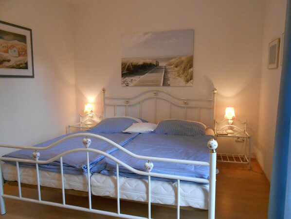 A bed with a metal frame, two bedside tables with lamps, a painting above the bed, and a small shelf on the wall.