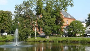 A building with a fountain in front, surrounded by trees and a pond. - Ankerplatz Vacation Rental - - Ankerplatz Vacation Rental (Brodersby)