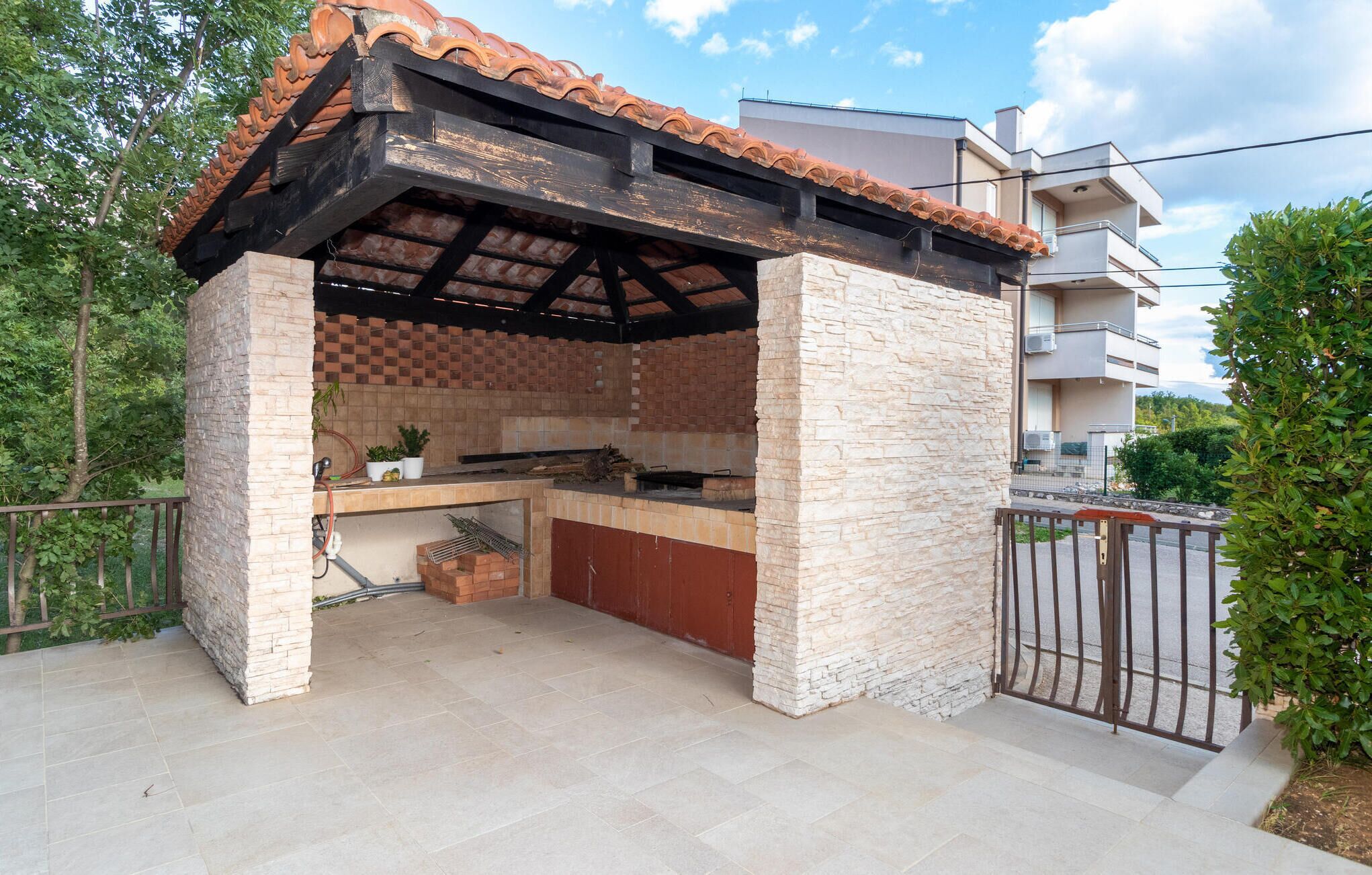 An outdoor kitchen with a tiled roof and stone walls, surrounded by a metal fence and greenery.