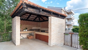 An outdoor kitchen with a tiled roof and stone walls, surrounded by a metal fence and greenery.