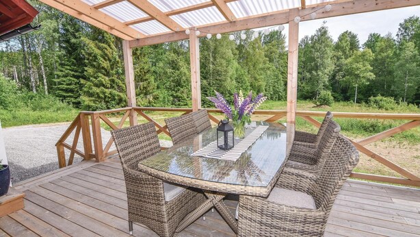 A covered outdoor seating area with a glass table and wicker chairs, surrounded by a wooden deck and a metal railing, overlooking a forested area.