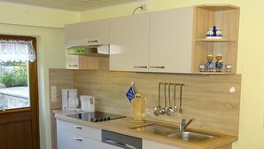 A compact kitchen with white cabinets, a built-in dishwasher, a sink, and a coffee maker. There is a small shelf with decorative items and a window with a lace curtain.
