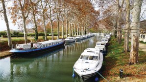 Un canal bordeado de barcos amarrados a la orilla, rodeado de árboles y una ribera cubierta de césped.
