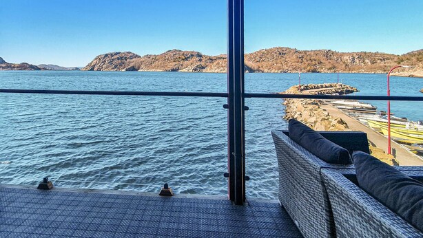 A view from a boat deck with a railing, a wicker sofa with cushions, and a rocky shoreline in the background.