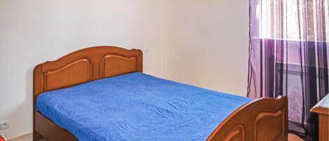 A single bed with a wooden headboard in a room with a plain wall and a window with sheer curtains.