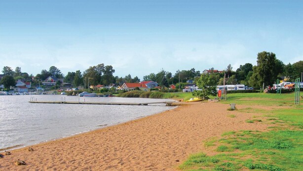 En strand med en træbro, der strækker sig ud i roligt vand, omgivet af huse og grøn vegetation.