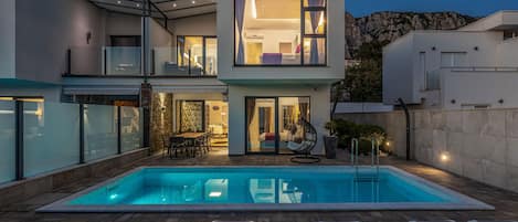 A modern two-story house with a swimming pool, outdoor seating, and a view of the surrounding landscape.