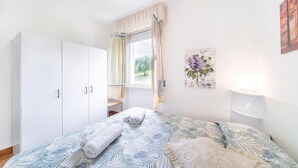 Et soveværelse med seng, skab, et vindue med gardiner, en vægmonteret lampe og et billede.