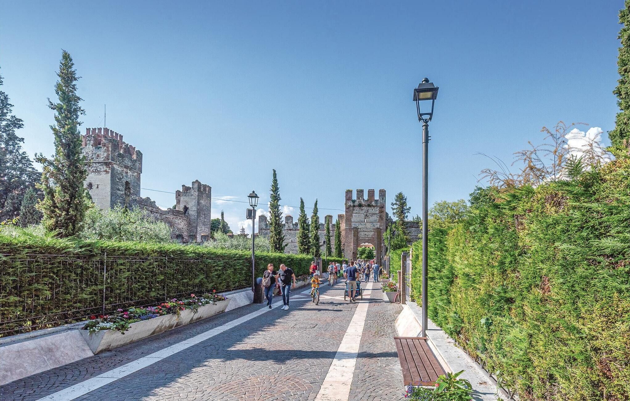 Uma passagem de pedras irregulares leva a um castelo histórico, com muralhas e torres de pedra, rodeado por vegetação e um lampião.