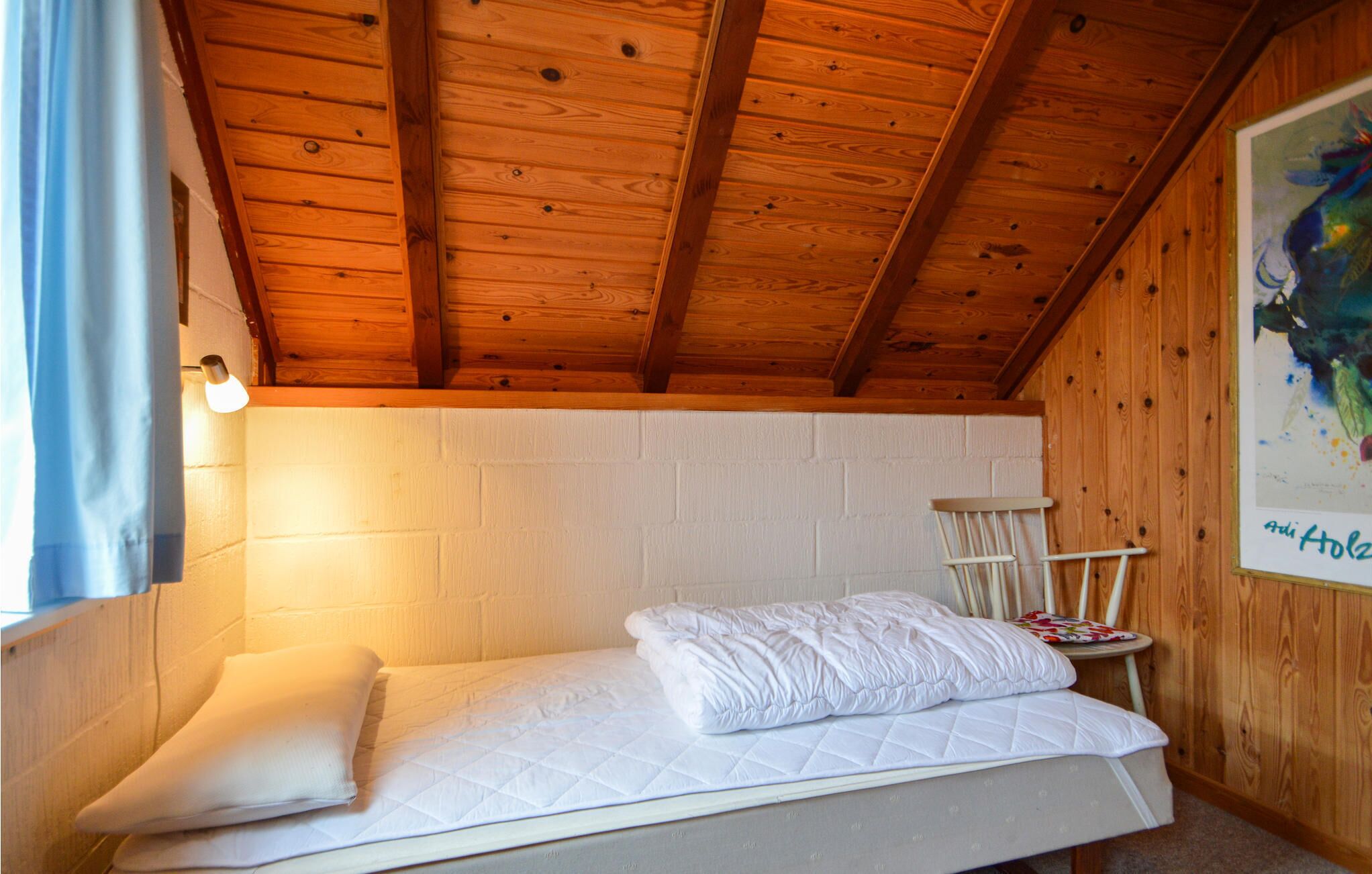 Een enkel bed met een gewatteerd matras en kussens, een houten plafond, een witte bakstenen muur en een ingelijste afbeelding aan de muur.