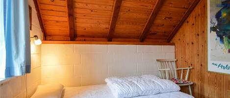Een enkel bed met een gewatteerd matras en kussens, een houten plafond, een witte bakstenen muur en een ingelijste afbeelding aan de muur.