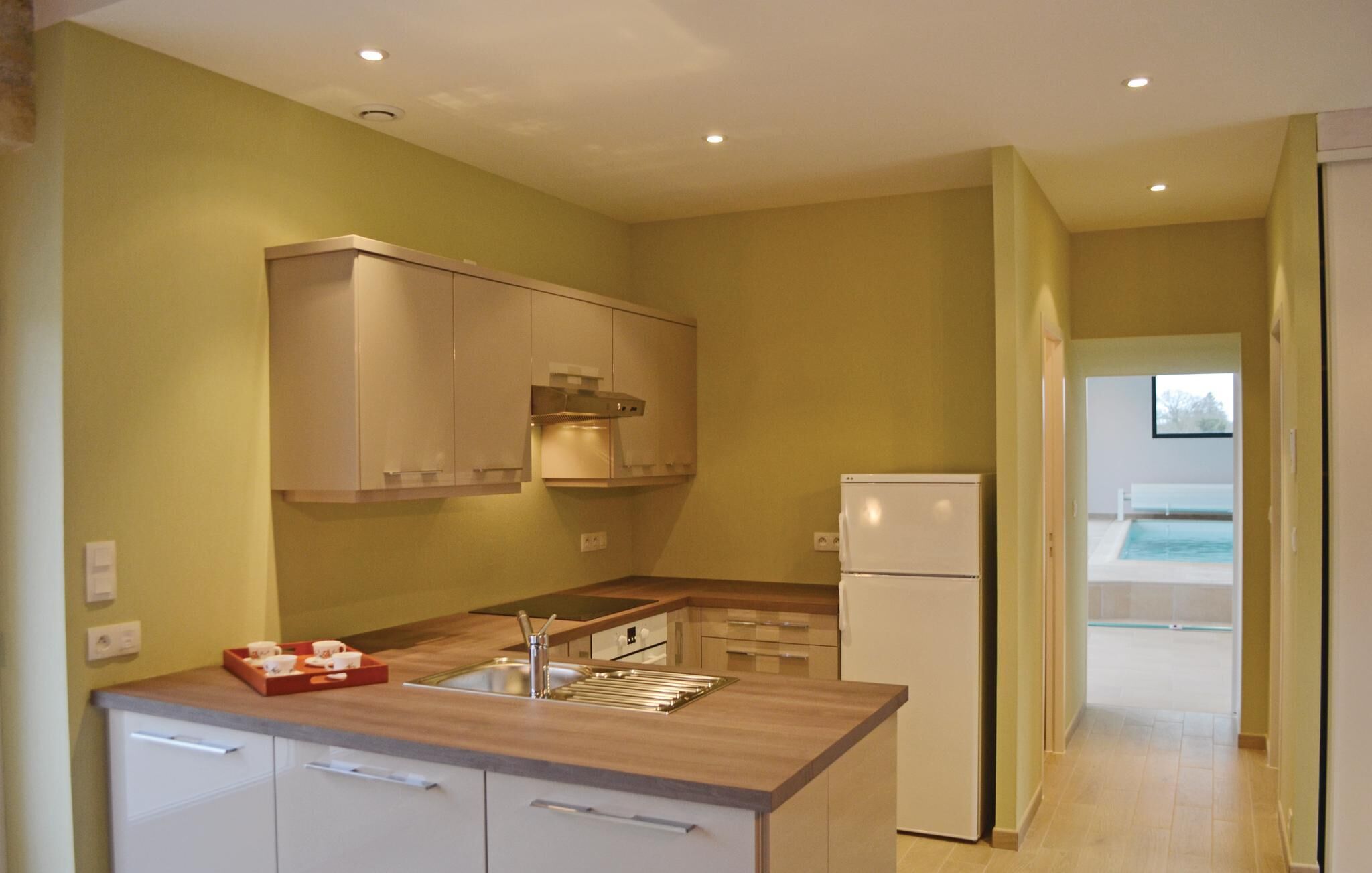 Eine moderne Küche mit weißen Schränken, einem eingebauten Ofen und einem Kühlschrank. Eine kleine Kochinsel mit Spüle und einer Tablett voll Tassen. Die Küche geht über in einen Bereich mit einem Pool und einer Glastür.