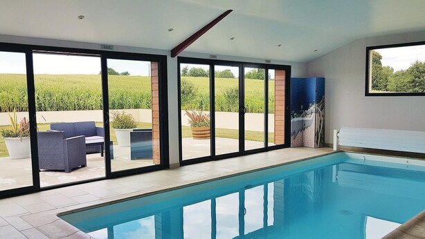 Une piscine intérieure moderne avec des parois en verre transparent, un sol carrelé et une vue sur un champ verdoyant.