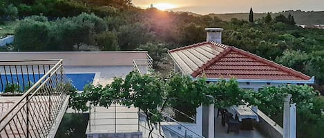 A residential building with a red-tiled roof, a stone staircase leading to a covered area, and a view of the sunset over a hilly landscape.