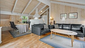 A spacious living area with a wooden ceiling, a black leather sofa, a dining table with chairs, and a kitchen in the background. - 3 bedroom stunning home in Glesborg (Glesborg)