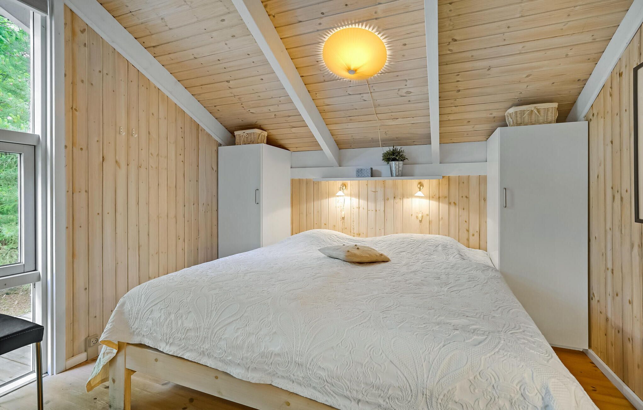 Une chambre avec un plafond en bois, un lit recouvert d’une couette blanche à texture, deux lampes de chevet et une fenêtre donnant sur un paysage verdoyant.