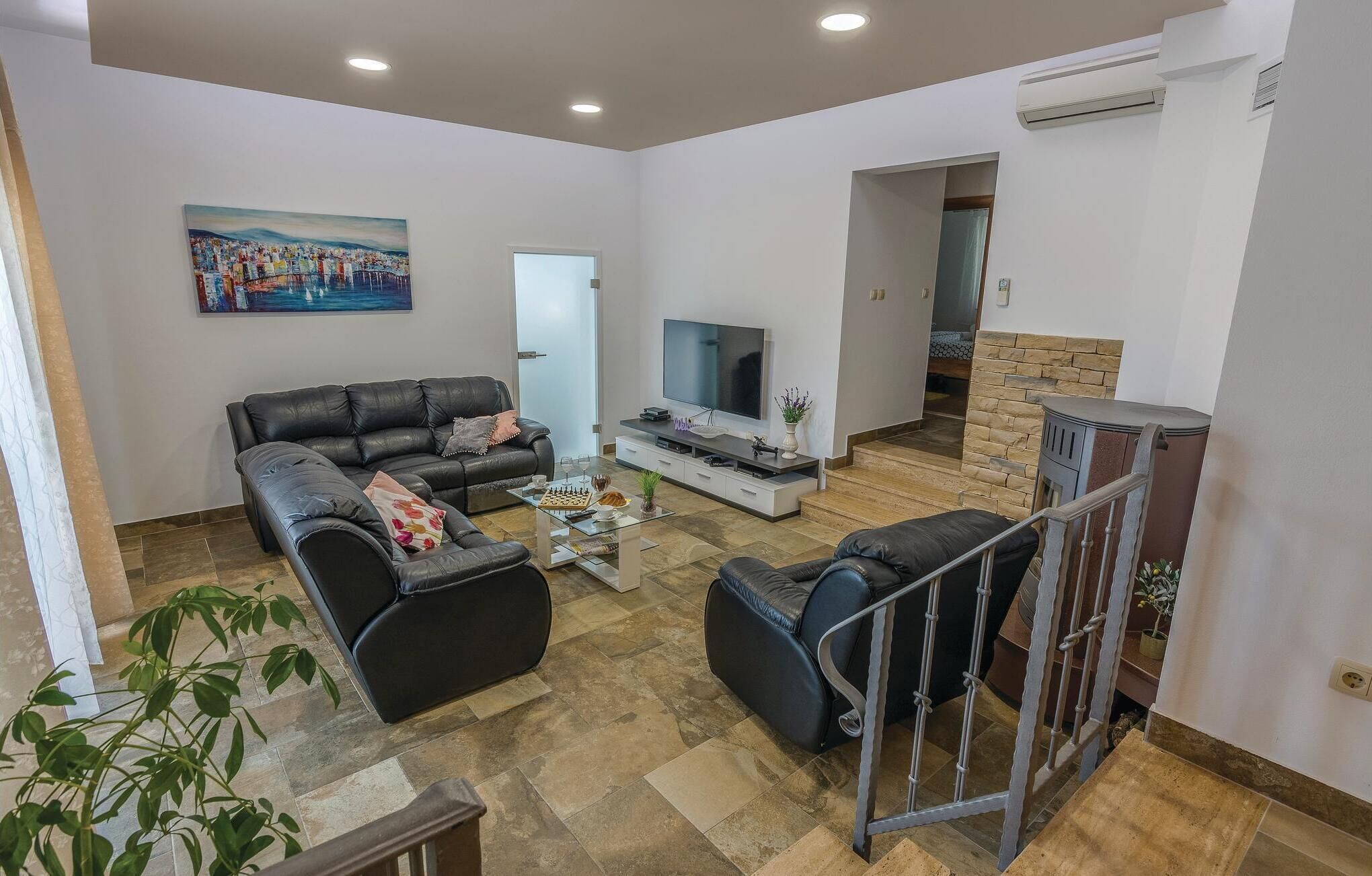 Ein geräumiges Wohnzimmer mit einer schwarzen Ledersofa, einem Glastisch und einem Flachbildfernseher. An der Wand hängt ein Gemälde und eine Treppe führt in einen anderen Raum.