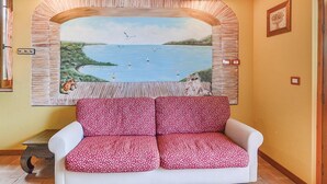 Un canapé blanc orné de coussins à motifs floraux rouges, dans une pièce dont un mur est décoré d’une fresque représentant un lac et des voiliers.