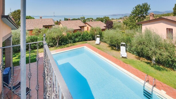 Une piscine rectangulaire à l’eau d’un bleu limpide, entourée d’une terrasse carrelée de couleur rouge. Deux chaises longues blanches sont disposées sur la terrasse, et la piscine est bordée d’une clôture métallique. Au fond, on aperçoit des maisons aux toits recouverts de tuiles rouges et une vue sur la mer.