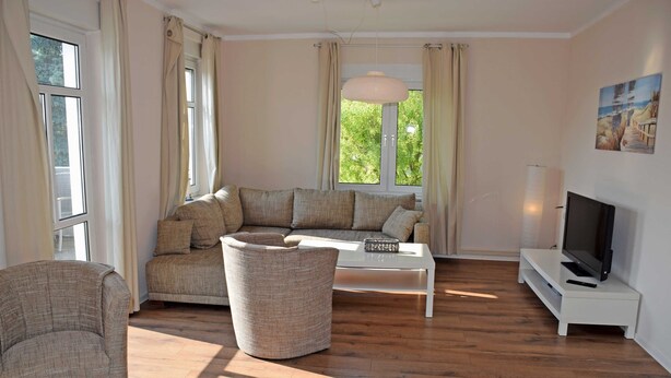A living room with a sofa, a coffee table, a TV on a stand, and a large window with curtains. - Vacation Rental in the Baltic Sea Resort of Göhren (01) - Vacation Rental with a Balcony (01) (Göhren)