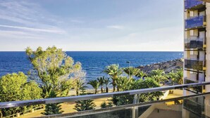 Un balcón con vista al mar y a la costa rocosa, rodeado de vegetación y un edificio moderno.