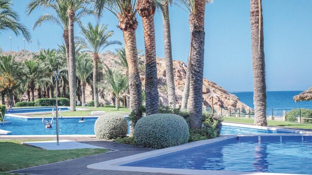 Zona de piscina con palmeras, vista despejada al mar y un promontorio rocoso al fondo.