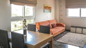 Una sala de estar con un sofá, una mesa y un florero con flores. Hay ventanas con persianas y un cuadro en la pared.