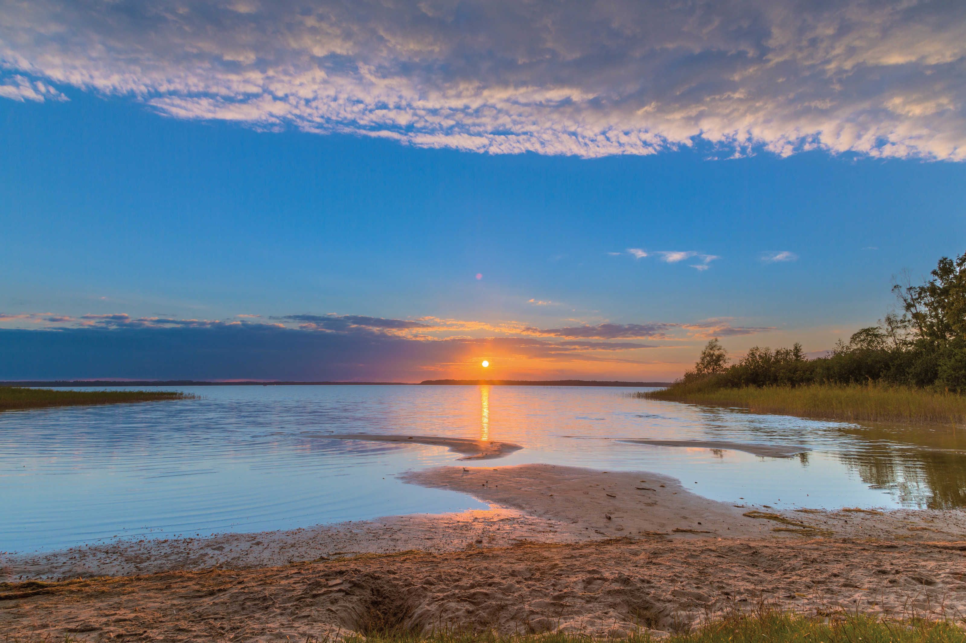 A sunset over a calm body of water with a sandy shore and sparse vegetation.