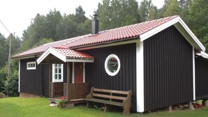 Une petite maison de plain-pied, avec un toit recouvert de tuiles rouges, un porche à l’avant doté d’un banc et une fenêtre ronde sur le côté.