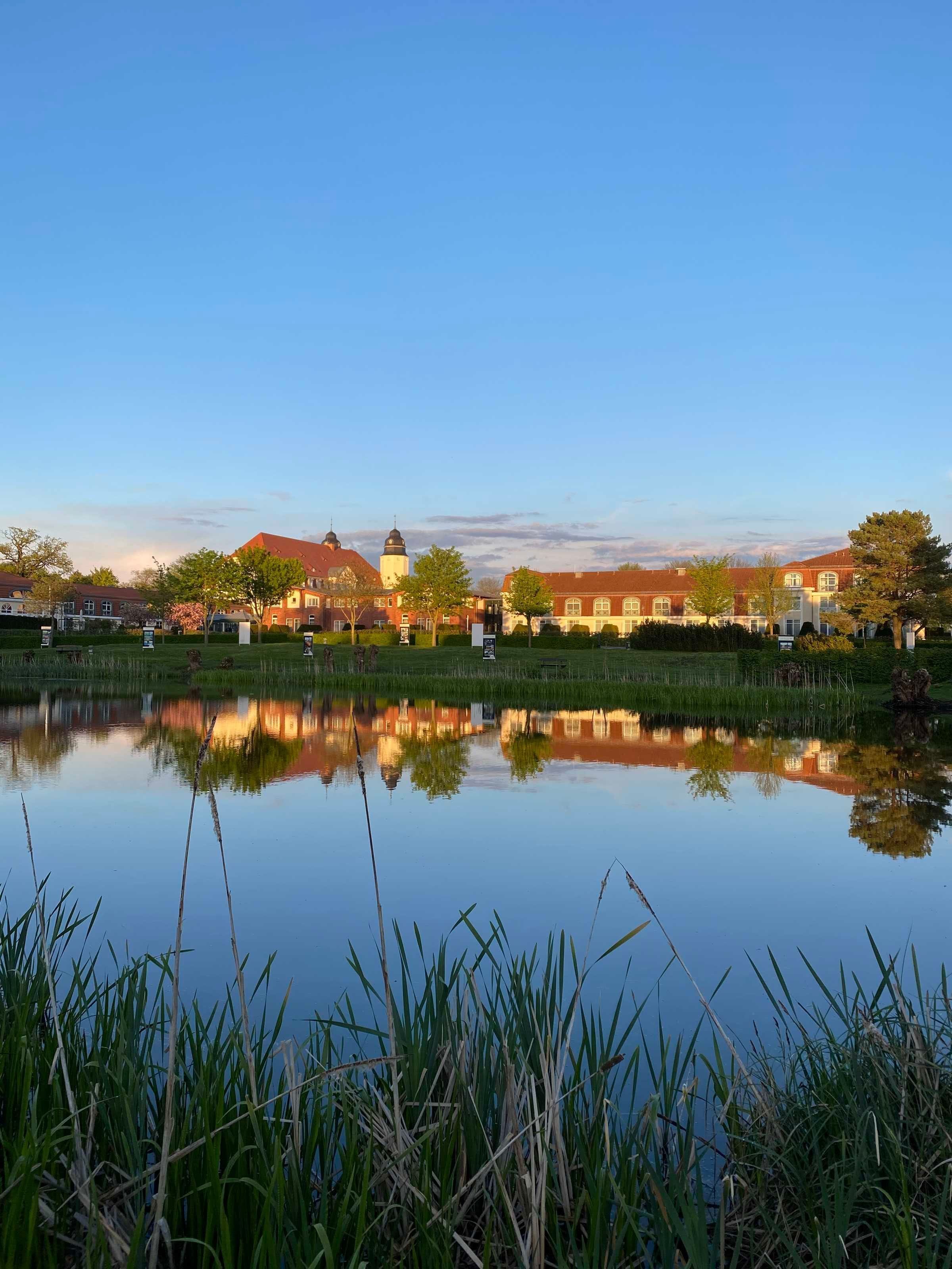 A serene lakeside view with a building featuring a prominent tower and dome, surrounded by lush greenery and a clear sky.