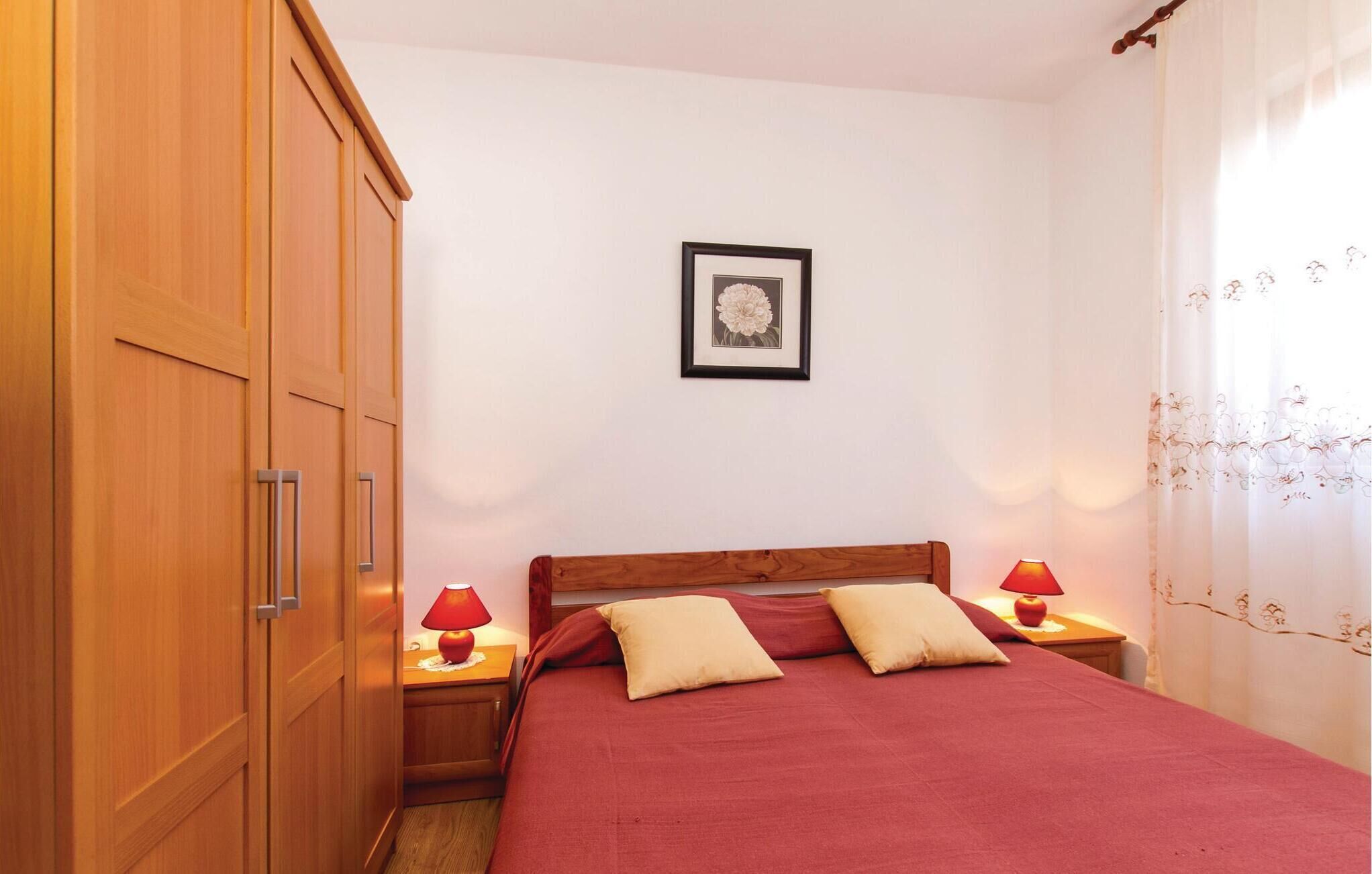 Une chambre avec une armoire en bois, un lit avec une literie rouge, deux lampes de chevet et un tableau encadré au mur.