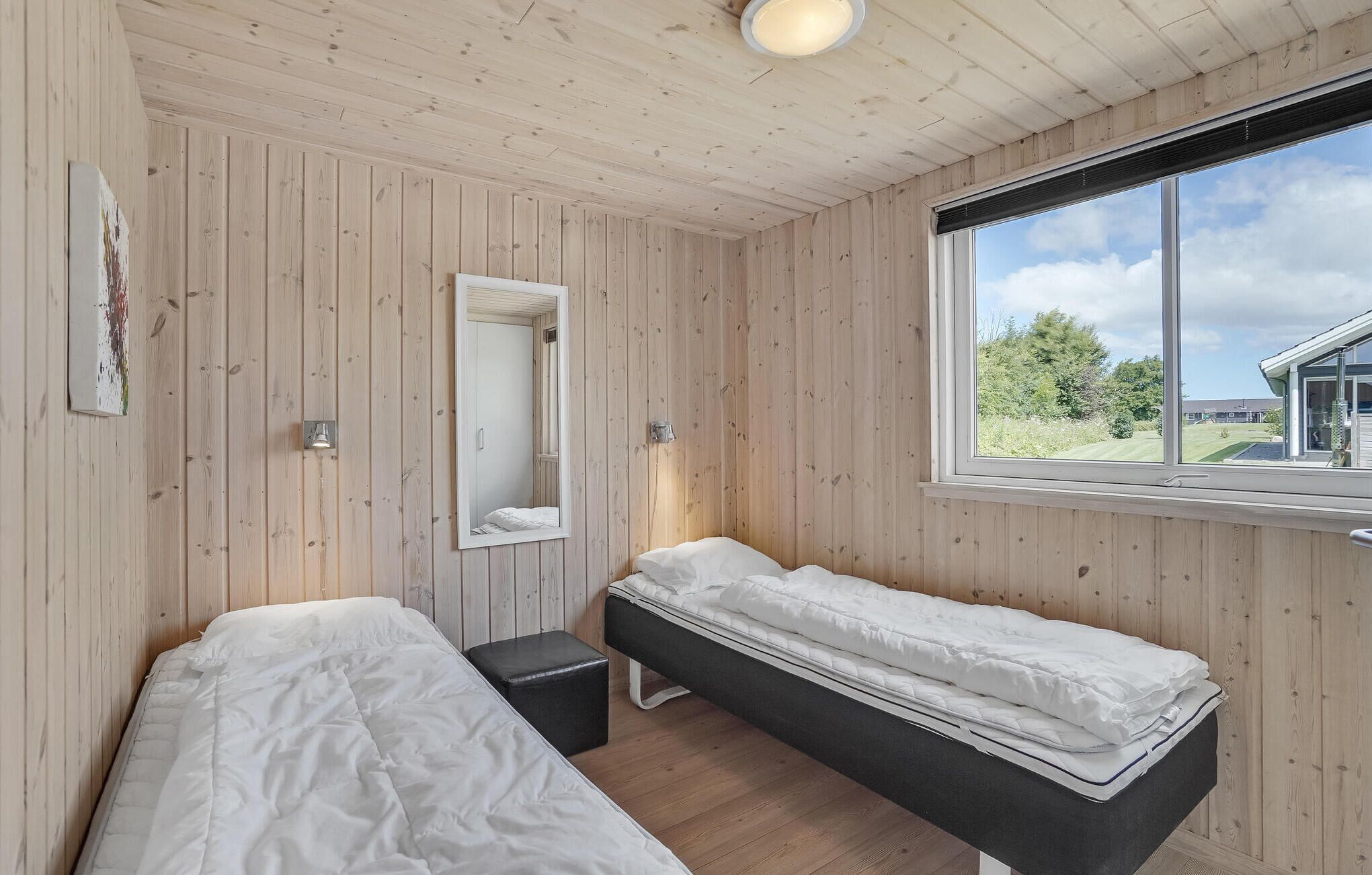Een kamer met houten lambrisering, een groot raam met uitzicht op de omgeving, een bed, een nachtkastje en een spiegel.