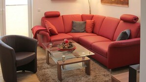 Un salon avec un canapé rouge, une table basse en verre, un fauteuil sombre et une étagère en bois ornée d’objets décoratifs.