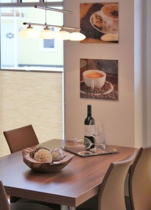 A dining area with a wooden table, chairs, a bowl of snacks, a wine bottle, and two framed pictures of coffee cups.