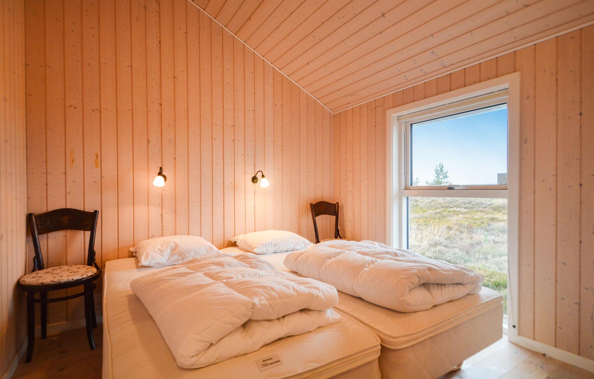 Un dormitorio acogedor con paredes de madera, una cama grande con ropa blanca, dos lámparas de pared y una ventana que ofrece vistas al exterior.