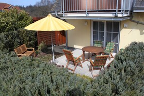Um pátio externo com mesa e cadeiras de madeira, um guarda-sol amarelo e um banco de madeira.