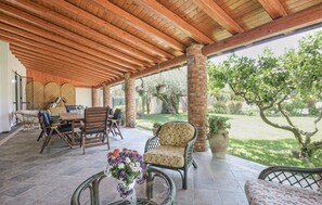 Ein überdachter Patio-Bereich mit Holzboden, Gartenmöbeln und Blick auf einen Garten mit Bäumen und Rasen.