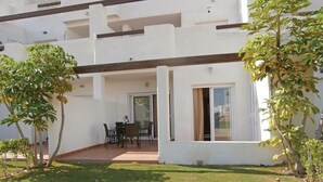 A modern building with a covered patio area, a dining table with chairs, and a small garden with trees. - Amazing apartment in Roldán with swimming pool (Roldán)
