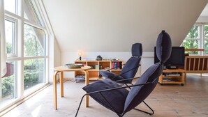Un loft moderne avec de hauts plafonds, de grandes fenêtres, un coin salon confortable avec un téléviseur, et une table en bois agrémentée de livres et d’une lampe.