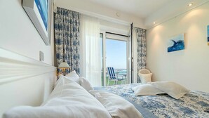 Hotellihuone, jossa on sänky, tuoli, pöytä ja näkymä merelle.
