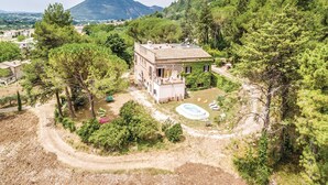 A two-story house with a pool and a tennis court, surrounded by dense forest and a mountain in the background.