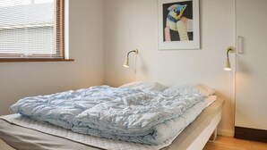 Un letto rifatto con un piumone azzurro chiaro e un copripiumino bianco trapuntato, due lampade da comodino, un quadro astratto incorniciato appeso alla parete e una finestra con persiane.