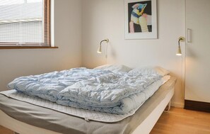 Siististi pedattu sänky, jossa on vaaleansininen peitto ja valkoinen, kuviollinen päällinen, kaksi yöpöytävalaisinta, seinällä oleva kehystetty abstrakti maalaus ja ikkuna, jossa on sälekaihtimet.