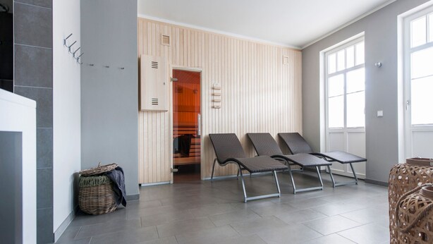 Ein modernes Hotelzimmer mit grau gefliestem Boden, einer Wand aus Holzpaneelen und einem großen Fenster. Es gibt zwei Sessel und einen Korbwäscher.