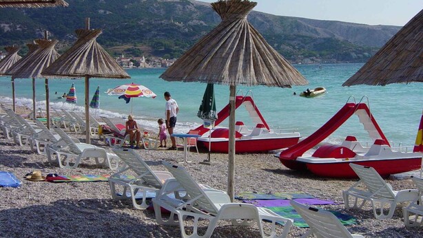 Una playa con filas de sillas blancas para tomar el sol, sombrillas de paja y un bote rojo.