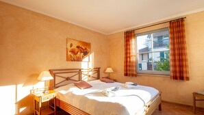 Une chambre à coucher avec un lit, des tables de chevet, une fenêtre avec des rideaux, un tableau au mur et une vue sur l’extérieur.