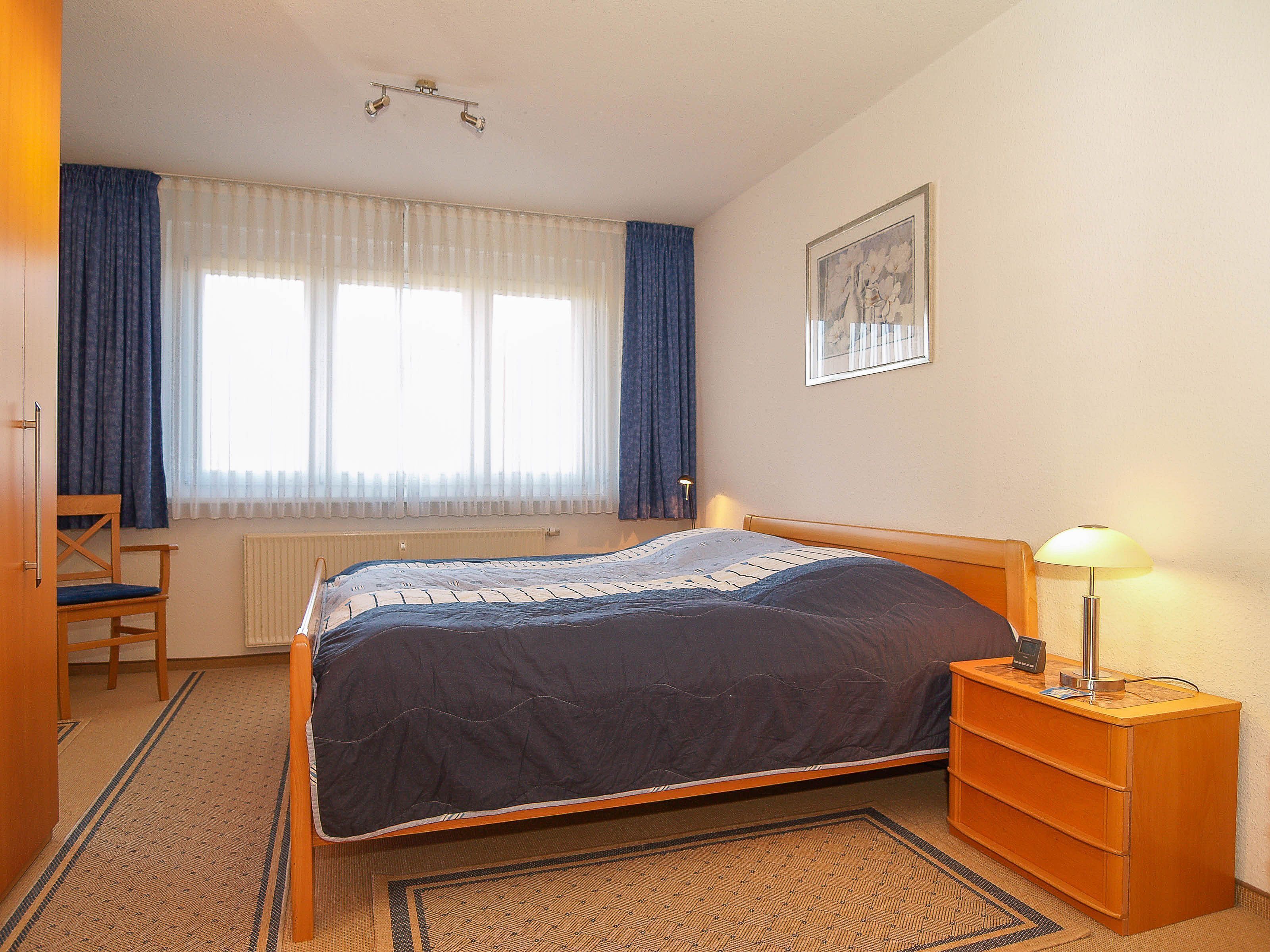 Ein Schlafzimmer mit Bett, Sessel, Nachttisch mit Lampe, Heizkörper und Fenster mit Vorhängen.