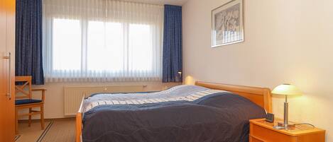 A bedroom with a bed, a chair, a bedside table with a lamp, a radiator, and a window with curtains.