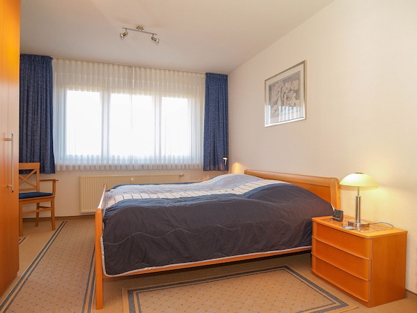 Ein Schlafzimmer mit Bett, Sessel, Nachttisch mit Lampe, Heizkörper und Fenster mit Vorhängen.